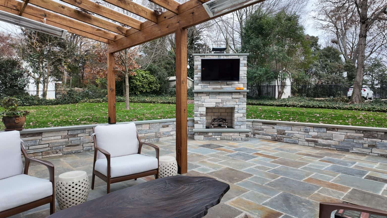 Stone patio under a wood pergola with an outdoor fireplace and mounted TV
